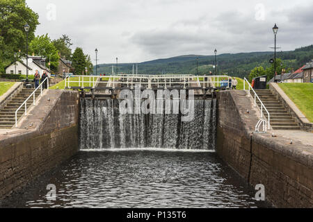 Fort Augustus, Scozia - Giugno 11, 2012: serratura con acqua che fluisce oltre il chiuso marrone scuro porte sul fiume Oich Canal mostra erba verde e case bianche Foto Stock