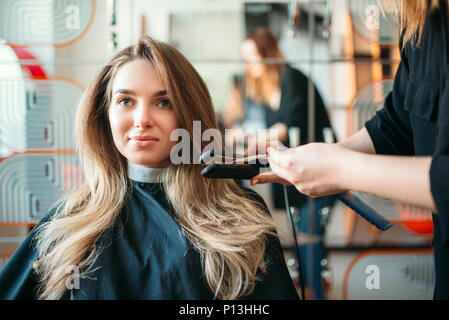 Parrucchiere raddrizza i capelli con un ferro da stiro, femmina client in un salone di parrucchiere. Acconciatura rendendo in studio di bellezza, parrucchiere facendo pettinatura Foto Stock