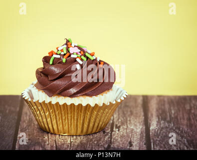 Tortina decorate con cremosa glassa di cioccolato e spruzza su tavola in legno rustico contro sfondo giallo Foto Stock