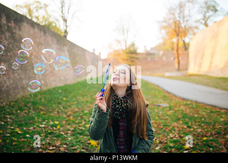 Attraente giovane donna sta godendo di rendere un bolle nel parco pubblico. Suolo coperto di foglie di autunno. Foto Stock