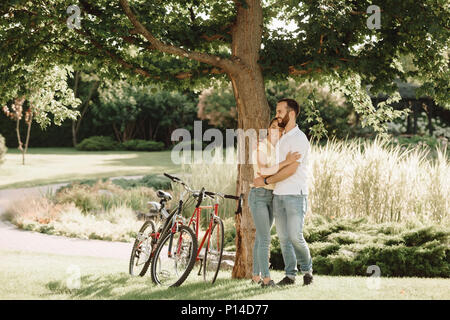 Giovane coppia romantica è costeggiata all'esterno. Una giovane coppia in amore in piedi sotto un albero e avvolgente. Data romantico nel parco. Foto Stock