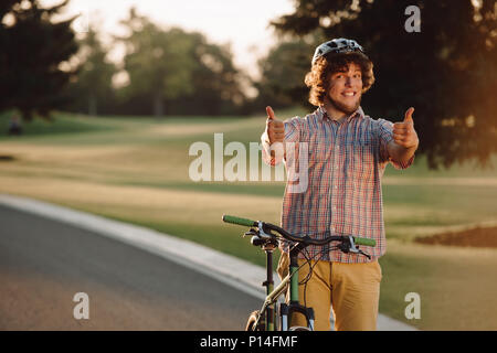 Felice ciclista dando due pollici fino all'esterno. Ritratto di Allegro ragazzo con moto gesticolando Thumbs up. Attivi in estate il tempo libero. Godetevi la vostra vita. Foto Stock