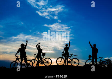 Sagome di giovani ciclisti al tramonto. Maschio e femmina di persone con biciclette a sventolare con le mani sul cielo della sera sfondo. Godendo l'infinito. Foto Stock