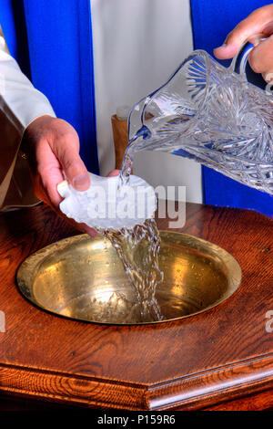 Un sacerdote (clero /ministro) versa acqua in un fonte battesimale. Foto Stock