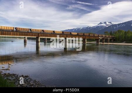 Treno merci Canadian Pacific Railway che attraversa la Columbia River Bank Water Revelstoke BC Canada. Panoramica panoramica delle Montagne Rocciose di Selkirk con vista panoramica sullo skyline Foto Stock