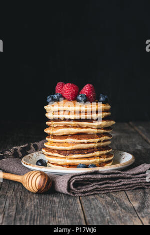 Pila di frittelle con miele e frutti di bosco freschi sul tavolo di legno su sfondo scuro. Messa a fuoco selettiva, copia spazio per il testo Foto Stock