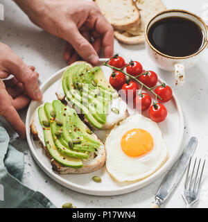 Toast con avocado, Uovo fritto, pomodori e tazza di caffè. Una sana prima colazione. Mani tenendo la piastra con una sana prima colazione il cibo. Concetto di vita sana Foto Stock