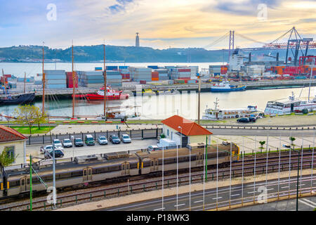 Il treno sulla Ferrovia dal porto commerciale con navi e contenitori, 25 de Abril ponte sul fiume Tago al tramonto, Cristo Re monumento sullo sfondo Foto Stock