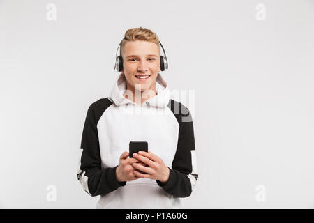 Ritratto di ragazzo adolescente 16-18 anni indossando hoodie sorridente e ascolto di musica sullo smartphone tramite cuffie senza fili isolati su bianco backgr Foto Stock