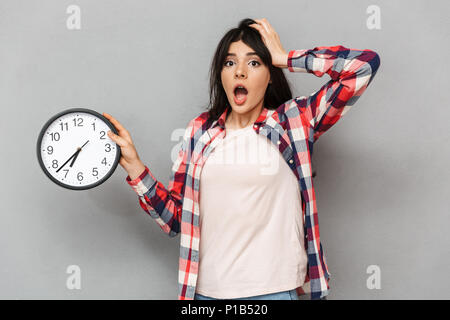 Immagine del sistema nervoso sconvolto giovane donna in piedi isolato su sfondo grigio parete orologio di contenimento. Foto Stock