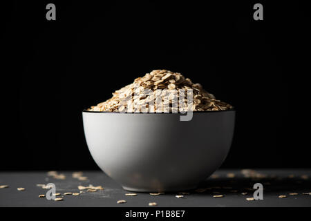 Primo piano di un bianco di ceramica ciotola piena di fiocchi d'avena su una tabella grigia, contro uno sfondo nero Foto Stock