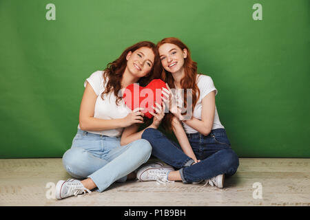 Ritratto di due attraenti redhead ragazze 20s in jeans seduto sul pavimento con le gambe incrociate e azienda carta rossa cuore isolato su sfondo verde Foto Stock