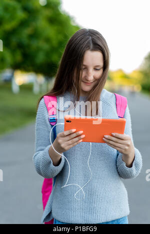 Bambina schoolgirl bruna. In estate la natura. Dopo le lezioni, egli mantiene le sue mani e tablet ascolta per te la musica nelle cuffie. Guardando il video. Sorridenti comunica in reti sociali. Foto Stock