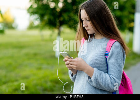 Bambina schoolgirl bruna. Dopo la scuola a scuola. Dietro la schiena è una rosa nello zaino. Nelle mani tiene uno smartphone. Ascolta la musica le cuffie guardare film. Scrive il messaggio ai genitori gli amici. Foto Stock