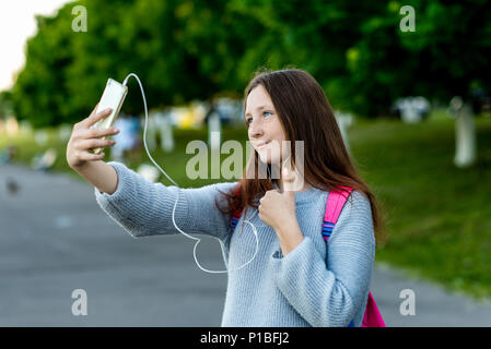 Bellissima bambina bruna schoolgirl. Talk su video chiamata sul telefono. Riprodurre la musica nelle cuffie. Fa un gesto con un gesto della mano che mostra un pollice in su. Comunicazione in reti sociali. Foto Stock