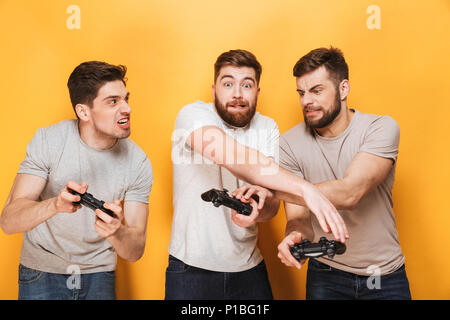 Tre giovani uomini divertenti holding joystick durante la riproduzione di video giochi isolate su sfondo giallo Foto Stock