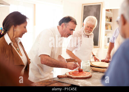 Senior gli amici a guardare lo chef diffusione di salsa marinara sulla pasta della pizza nella classe di cucina Foto Stock