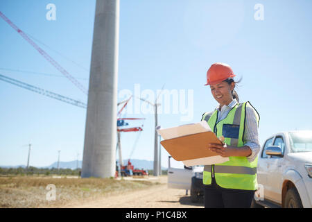 Sorridente ingegnere femmina con blueprint a sunny turbina eolica impianto di alimentazione Foto Stock