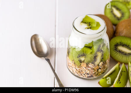 Yogurt bianco con muesli in recipiente di vetro con pezzi di kiwi sulla parte superiore e interi pezzi sul lato destro. Foto Stock