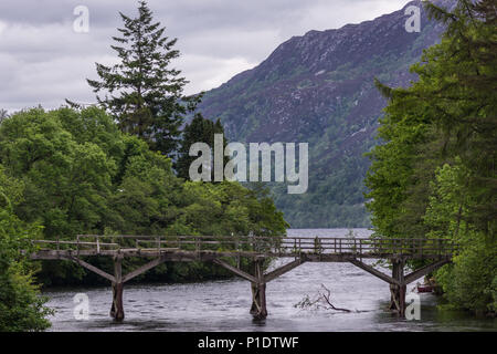 Fort Augustus, Scozia - Giugno 11, 2012: Vista dettagliata del danneggiato piede legno ponte sul fiume Oich. Cintura verde tra argento e acqua pesante cloudsc grigio Foto Stock