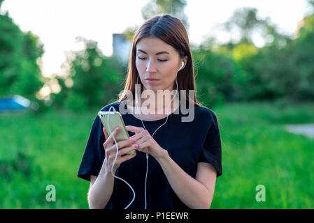 Bella bruna ragazza in estate nel parco. Al di fuori della città in natura. Tiene le mani del telefono. Ascolta le cuffie. Scrive i messaggi sulle reti sociali. Sullo smartphone è la visione di un filmato. Foto Stock