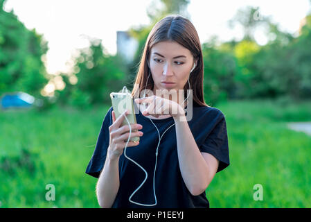 La bruna ragazza estate nel parco natura.sto guardando la. Tiene le mani con un telefono con le cuffie. Con il gesto della mano e del dito mostra. Gesti emotivo, mi aspetto dopo l. Foto Stock