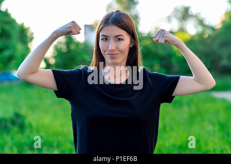 La bruna ragazza sorride felicemente. Estate in campo su un prato verde. Mani che mostra bicipiti. Le emozioni sono scherzi del potere femminile. Foto Stock