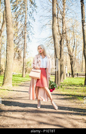 Moda ritratto di donna elegante in tessuto di colore rosa. La ragazza di setosa impermeabile, stivaletti, gonna e camicetta holding borsetta e fiori Foto Stock