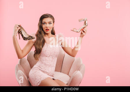 Immagine di perplesso shopping donna nel vestire la scelta e la holding diverse scarpe mentre è seduto in poltrona isolate su sfondo rosa Foto Stock