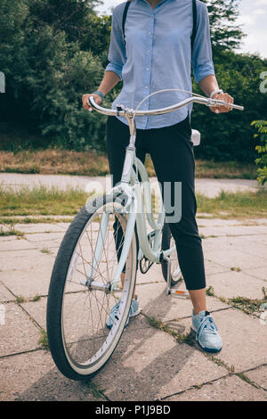 Irriconoscibile giovane femmina in abito casual in piedi accanto alla sua bici cruiser holding manubrio. Close-up di donna sulla bicicletta al giorno di sole. Foto Stock