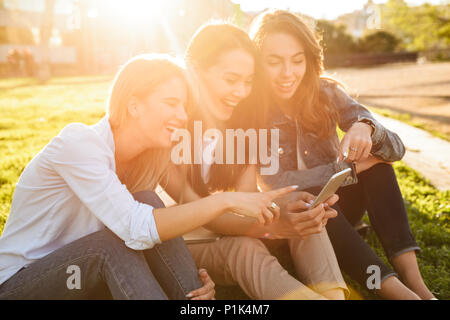 Foto di felice tre amici le donne sedute su erba di parcheggiare all'esterno guardando a parte utilizzando il telefono cellulare. Foto Stock