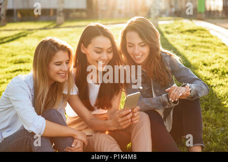 Foto di felice tre amici le donne sedute su erba di parcheggiare all'esterno guardando a parte utilizzando il telefono cellulare. Foto Stock