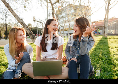 Foto di incredibile tre amici le donne sedute su erba di parcheggiare all'esterno guardando a parte utilizzando il computer portatile. Foto Stock