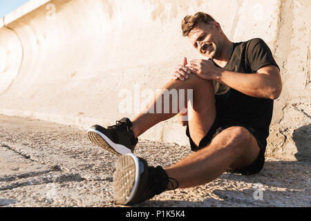 Ritratto di un ferito sportivo nel dolore che soffrono di dolore al ginocchio mentre seduti all'aperto Foto Stock