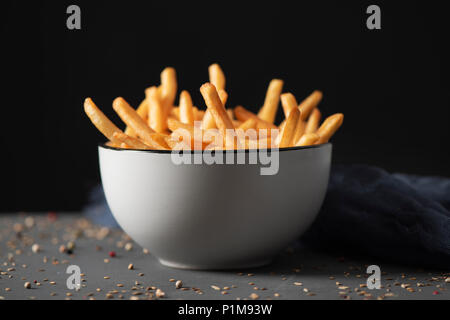 Alcuni appetitosi patatine fritte servite in un bianco di ceramica ciotola, collocato sul grigio di una tavola in legno rustico, contro uno sfondo nero Foto Stock