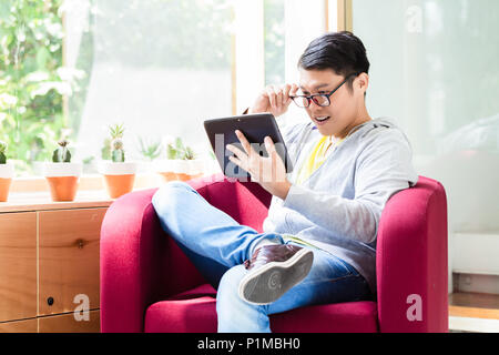 Giovane uomo asiatico sorpreso da online informazioni visualizzate su un tablet PC mentre è seduto in poltrona per interni Foto Stock