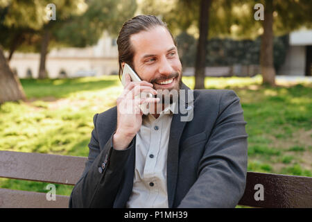 Foto di felice uomo barbuto 30s in abito formale avente conversazione mobile mentre è seduto sulla panchina nel parco della città durante la giornata di sole Foto Stock