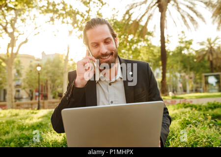 Foto Dettaglio di pragmatico uomo bello con capelli legati a lavorare sul notebook argento e avente mobile call mentre sedendo nel parco della città durante il sunny Foto Stock