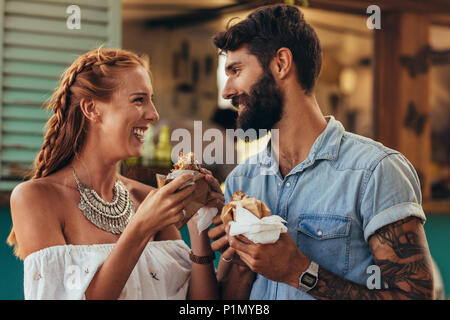 Ritratto di felice coppia giovane avente burger all'esterno. Giovane uomo e donna in piedi vicino a un carrello di cibo e mangiare hamburger. Foto Stock