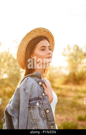 Foto di eleganti donna europea con lunghi capelli scuri che indossa cappello di paglia e la camicia di jeans cercando in disparte mentre passeggiate in campagna durante la giornata di sole Foto Stock