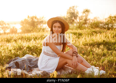 L'immagine orizzontale di bella bruna donna con lunghi capelli scuri che indossa cappello di paglia e abito bianco seduto sull'erba durante il riposo nella natura in estate Foto Stock