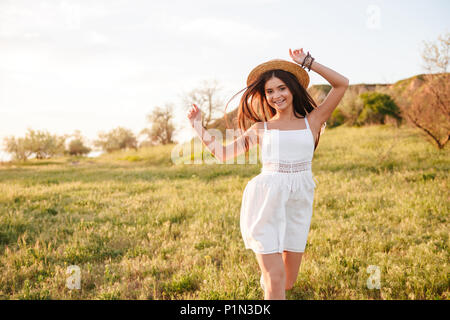 Immagine di allegro vivace da donna con lunghi capelli scuri che indossa cappello di paglia e abito bianco, sorridente e in esecuzione su erba in campagna durante il sole luminoso Foto Stock