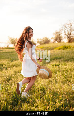 Foto di adorabile donna europea con lunghi capelli scuri che indossa cappello di paglia e abito bianco, camminando attraverso il prato in campagna durante la giornata di sole Foto Stock