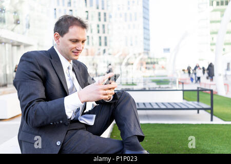 Giovane imprenditore sorridente seduto sulla panchina nel parco, utilizzando tenendo lo smartphone mobile telefono cellulare sorridente guardando giù texting in tuta e cravatta, chee Foto Stock