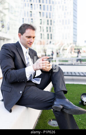 Giovane imprenditore seduto sulla panchina nel parco, utilizzando lo smartphone mobile telefono cellulare sorridente guardando giù texting in tuta e cravatta, allegro su intervie Foto Stock