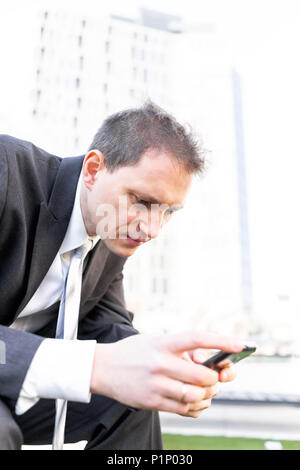 Giovane imprenditore seduto sulla panchina del parco in primo piano della faccia, utilizzando lo smartphone mobile telefono cellulare sorridente guardando giù texting in tuta e cravatta su in Foto Stock