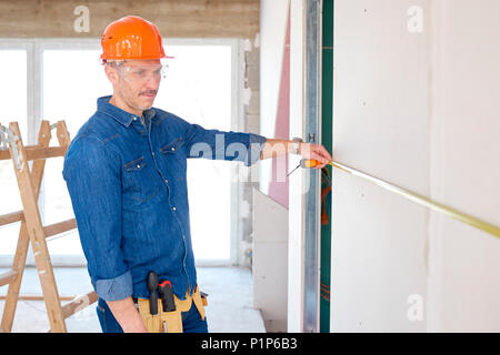 Il tuttofare utilizzando una misura di nastro mentre si lavora sul sito in costruzione. Repairman sicurezza indossare casco e occhiali di protezione. Foto Stock