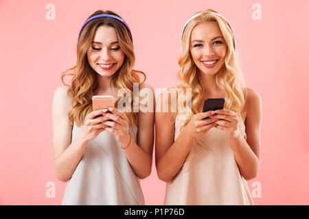 Due felici donne belle in pigiama e cuffie utilizzando gli smartphone mentre si ascolta la musica su sfondo rosa Foto Stock