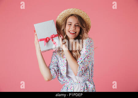 Ritratto di un soddisfatto giovane donna in abiti estivi e cappello di paglia azienda confezione regalo isolate su sfondo rosa Foto Stock