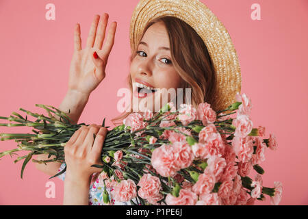 Close up ritratto di un bel giovane donna in abiti estivi e cappello di paglia tenendo i garofani bouquet e agitando con mano isolate su sfondo rosa Foto Stock